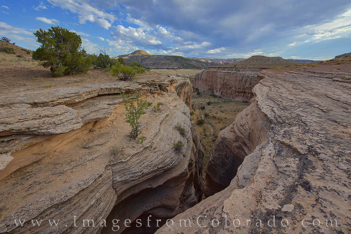 From the edges of a slot canyon, this photograph looks down into the red standstone walls. In the distance, the valley that holds...
